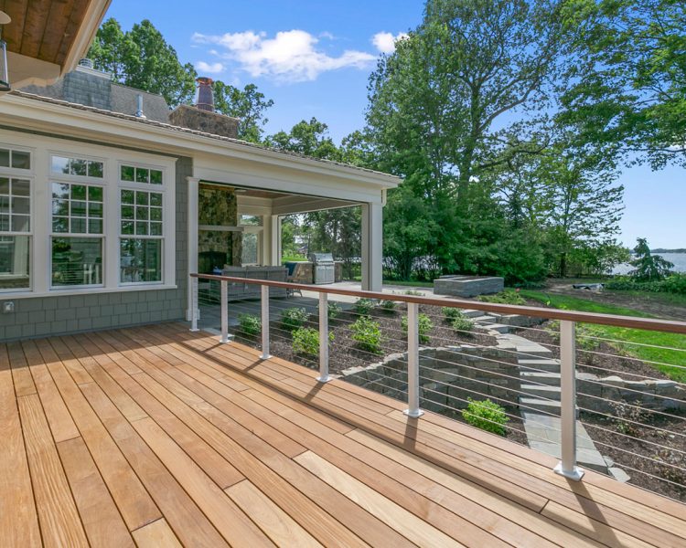 A spacious lakeside deck featuring a built-in outdoor kitchen and dining area, overlooking a serene lake surrounded by mature trees.