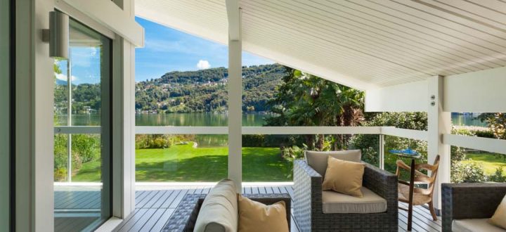 A peaceful lakeside deck area furnished with comfortable seating, covered by a white pergola, overlooking a serene lake surrounded by lush greenery.