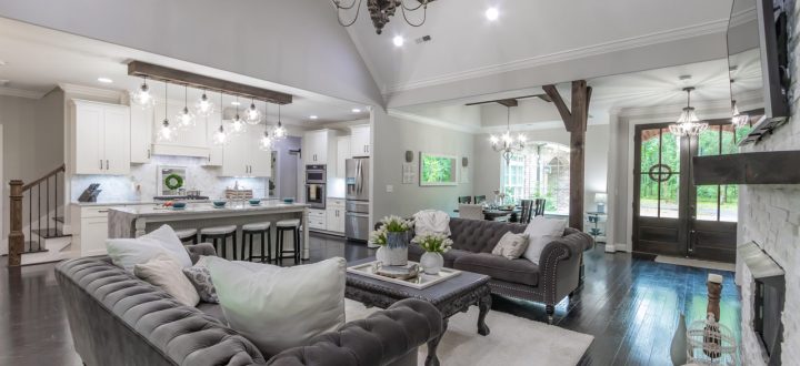 A spacious open plan interior showcasing a modern kitchen with white cabinetry and marble countertops adjacent to a living area with dark wood floors and luxurious grey furnishings.