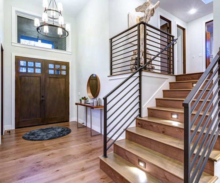 A contemporary home entryway featuring a wooden door with glass panels, modern staircase with metal balusters and wooden steps, under a chic chandelier.